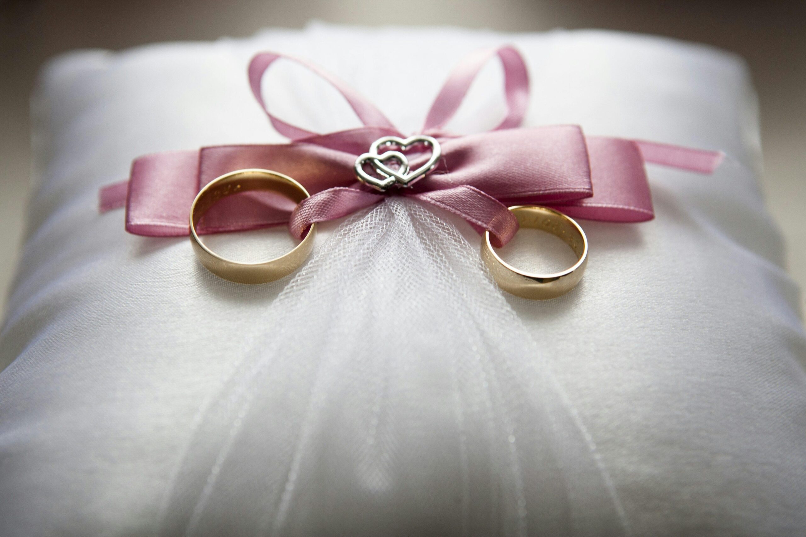 Close-up of elegant wedding rings on a silk pillow with a pink ribbon and a heart decoration, symbolizing love.