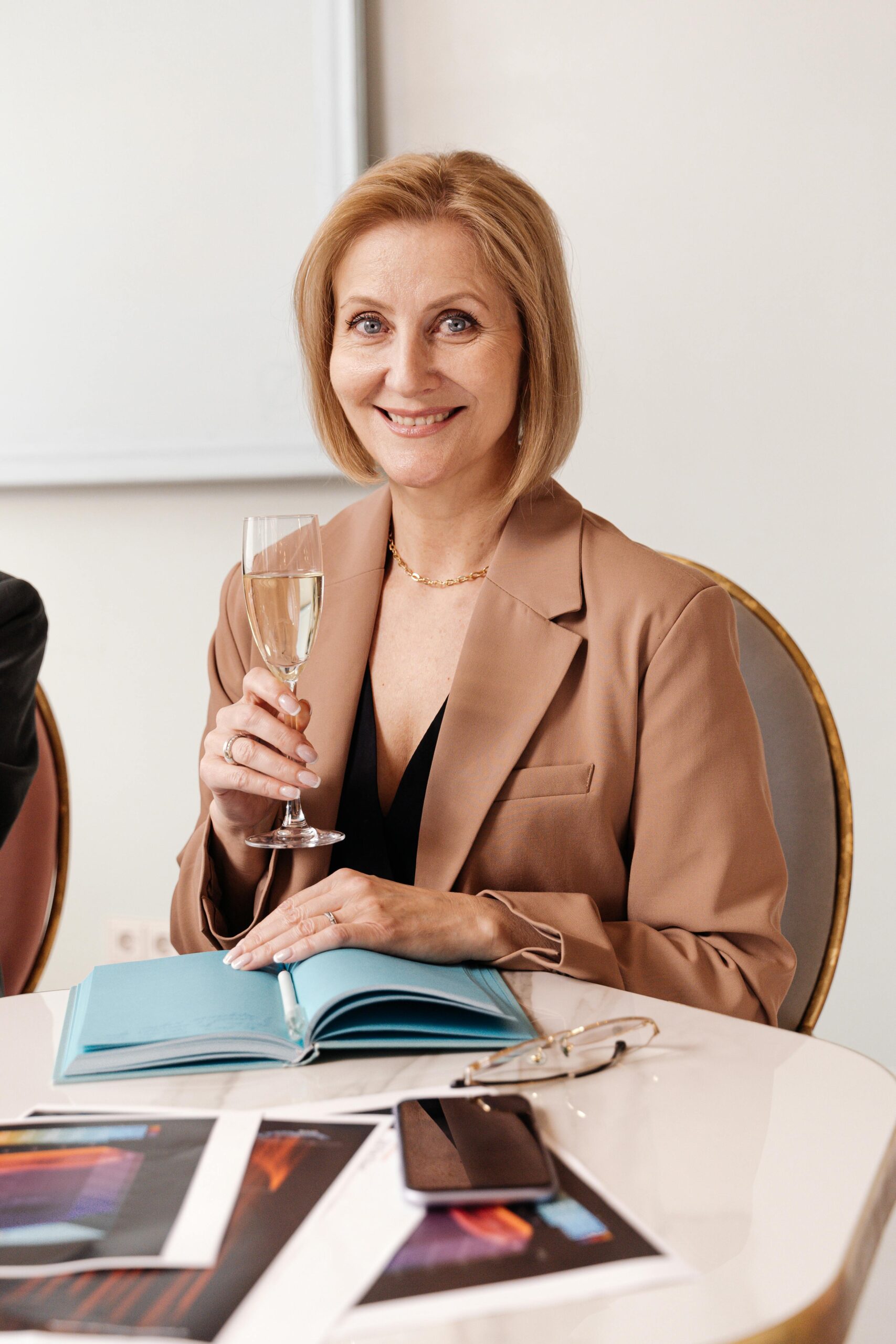 Smiling woman in a blazer holding a glass of wine during a business meeting indoors.